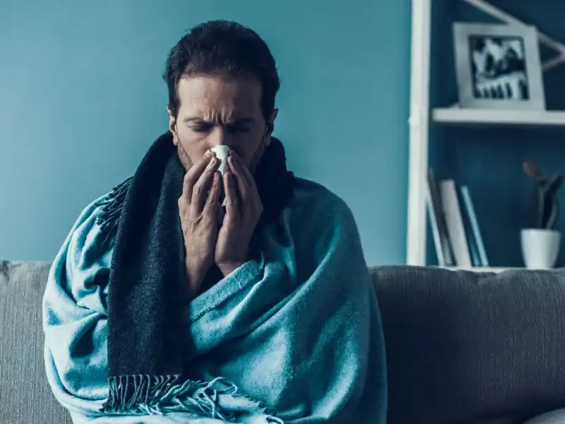 A sick man sneezing while covered in blankets on the couch.