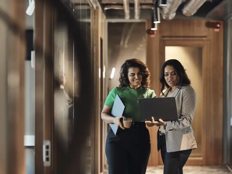 Two coworkers converse as they study a laptop computer while standing in an office hallway.