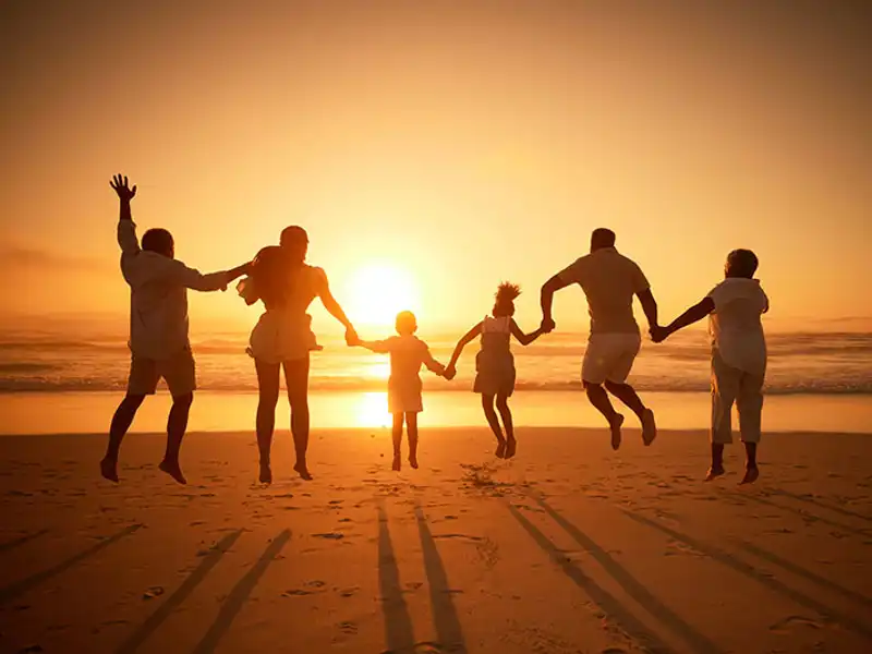 A silhouetted family holds hands and jumps into the air simultaneously on a sandy beach at sunset.