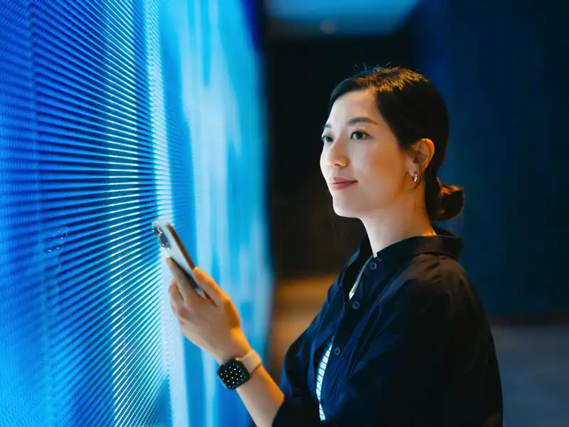 Woman holding phone standing next to a lighted wall.
